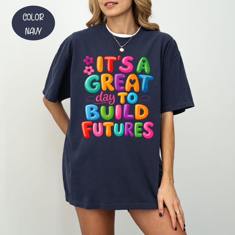 Pode incluir: Camiseta azul marinho com a frase "IT'S A GREAT day TO BUILD FUTURES" em letras coloridas e em bolhas. O design inclui uma flor e um cora&ccedil;&atilde;o. A camiseta &eacute; de estilo confort&aacute;vel e casual.