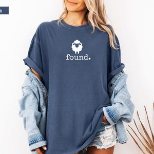 May include: Denim blue t-shirt with a white sheep graphic and the word "found." The shirt is a relaxed fit and has rolled-up sleeves. A denim jacket is worn over the shirt. The shirt is a casual style.