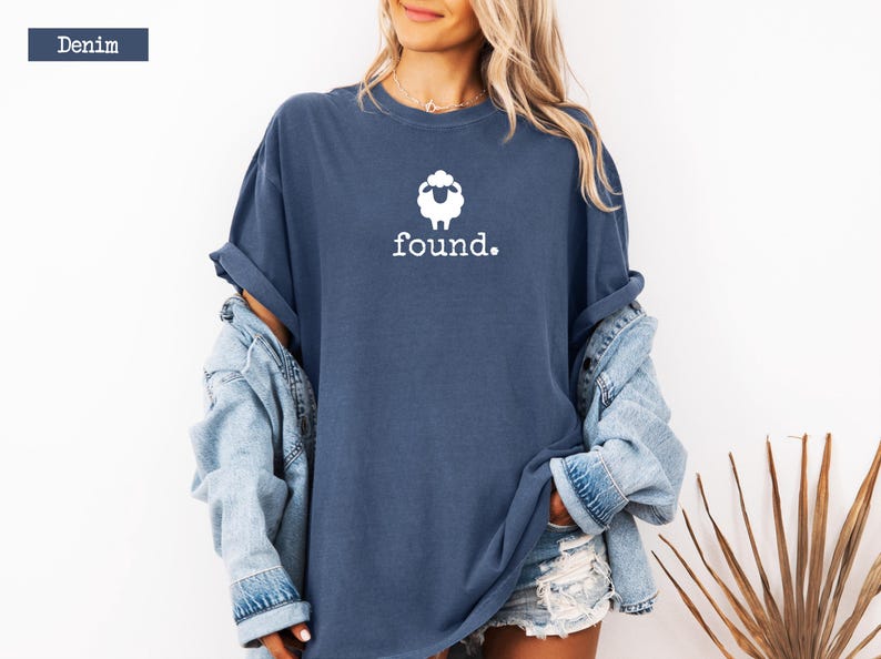 May include: Denim blue t-shirt with a white sheep graphic and the word "found." The shirt is a relaxed fit and has rolled-up sleeves. A denim jacket is worn over the shirt. The shirt is a casual style.