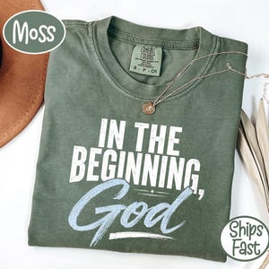 Peut inclure: Un t-shirt à manches courtes vert mousse avec une encolure ronde. Le vêtement présente le texte imprimé en blanc "IN THE BEGINNING, God" sur la poitrine. Un petit collier pendentif doré repose sur la zone du col du t-shirt.
