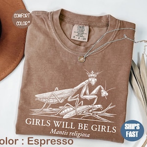 Peut inclure: T-shirt couleur expresso avec un motif blanc de deux mantes religieuses et le texte "GIRLS WILL BE GIRLS Mantis religiosa". Le t-shirt a un col rond et des manches courtes. Un collier argenté avec un pendentif est posé sur le t-shirt.
