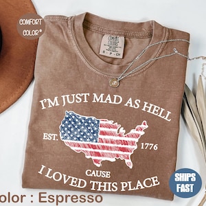 Peut inclure: T-shirt couleur expresso avec le texte "I'M JUST MAD AS HELL CAUSE I LOVED THIS PLACE" et un drapeau américain. Le t-shirt est de la marque Comfort Colors et a un col rond. Un collier argenté avec un pendentif est sur le t-shirt.