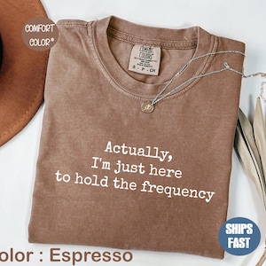 May include: Espresso-colored t-shirt with the text "Actually, I'm just here to hold the frequency." The shirt has a crew neck and a small tag at the neckline. A silver necklace with a pendant is draped across the shirt.