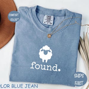May include: A blue jean Comfort Colors t-shirt with a white sheep graphic and the word "found." The shirt has a crew neck and short sleeves. A gold necklace with a pendant is draped across the shirt. The text "SHIPS FAST" is on a blue circle.