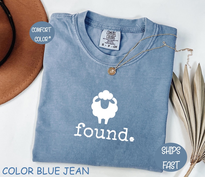 May include: A blue jean Comfort Colors t-shirt with a white sheep graphic and the word "found." The shirt has a crew neck and short sleeves. A gold necklace with a pendant is draped across the shirt. The text "SHIPS FAST" is on a blue circle.