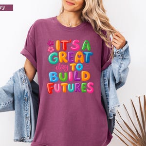 Pode incluir: Camiseta cor de berry com o texto "IT'S A GREAT day TO BUILD FUTURES" em letras de bolhas coloridas. A camiseta tem um corte folgado e mangas curtas. Uma jaqueta jeans est&aacute; sobre os ombros.