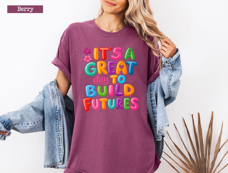 Pode incluir: Camiseta cor de berry com o texto "IT'S A GREAT day TO BUILD FUTURES" em letras de bolhas coloridas. A camiseta tem um corte folgado e mangas curtas. Uma jaqueta jeans est&aacute; sobre os ombros.