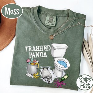 K&ouml;nnte beinhalten: Moosgr&uuml;nes T-Shirt mit dem Schriftzug "Trashed Panda" und einer Grafik eines Waschb&auml;ren, einer Toilette und eines M&uuml;lleimers. Das Shirt hat einen Rundhalsausschnitt und kurze &Auml;rmel. Eine goldene Halskette ist auf dem Shirt drapiert.