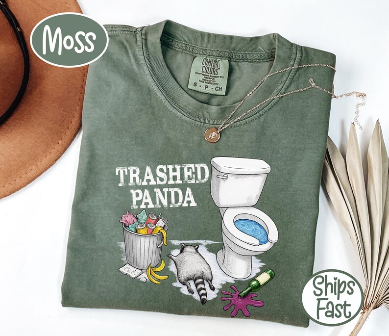 K&ouml;nnte beinhalten: Moosgr&uuml;nes T-Shirt mit dem Schriftzug "Trashed Panda" und einer Grafik eines Waschb&auml;ren, einer Toilette und eines M&uuml;lleimers. Das Shirt hat einen Rundhalsausschnitt und kurze &Auml;rmel. Eine goldene Halskette ist auf dem Shirt drapiert.