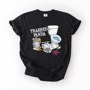 K&ouml;nnte beinhalten: Schwarzes T-Shirt mit dem Text "Trashed Panda" &uuml;ber einer Grafik eines Waschb&auml;ren, einer Toilette, einem M&uuml;lleimer und einer versch&uuml;tteten Flasche. Der M&uuml;lleimer ist mit bunten Gegenst&auml;nden gef&uuml;llt.