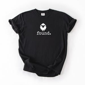May include: Black t-shirt with a white sheep graphic and the word "found." printed below. The sheep graphic is a simple outline with a fluffy head. The shirt is a basic crew neck.