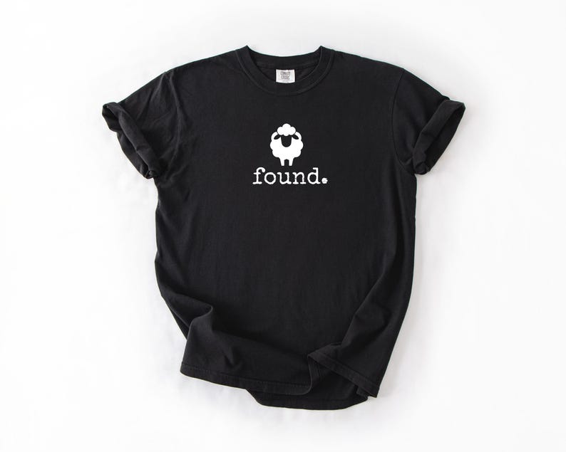 May include: Black t-shirt with a white sheep graphic and the word "found." printed below. The sheep graphic is a simple outline with a fluffy head. The shirt is a basic crew neck.