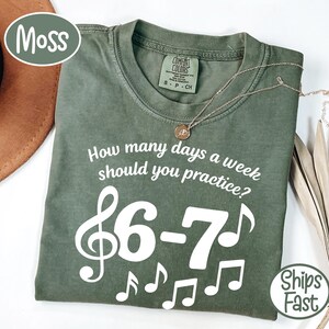 Peut inclure: Un t-shirt vert mousse avec le texte "How many days a week should you practice ?" et des notes de musique. Le t-shirt porte également le texte "6-7". Un collier en or avec un pendentif est sur le t-shirt.