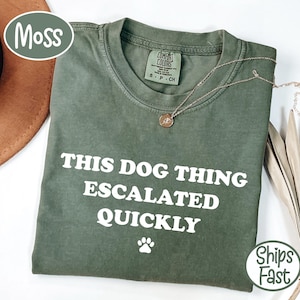 Puede incluir: Una camiseta de manga corta color verde musgo con cuello redondo. La prenda presenta el texto impreso en blanco "THIS DOG THING ESCALATED QUICKLY" seguido de un pequeño icono de huella blanca. Se ve una etiqueta de Comfort Colors en el cuello.