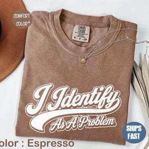 Könnte beinhalten: Ein espressofarbenes Kurzarm-T-Shirt mit Rundhalsausschnitt. Das Shirt ist mit einem weißen Aufdruck versehen, der den Text "I Identify As A Problem" in einer Schriftart im Retro-Stil zeigt. Das Innenetikett am Kragen trägt den Markennamen Comfort Colors.