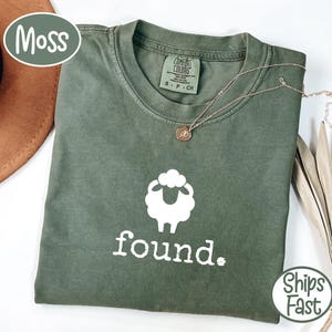 Op de afbeelding: Een mosgroen t-shirt met een witte schapenafbeelding en het woord "found" in het wit. Het shirt heeft een ronde hals en korte mouwen. Een gouden ketting met een kleine hanger ligt op het shirt.