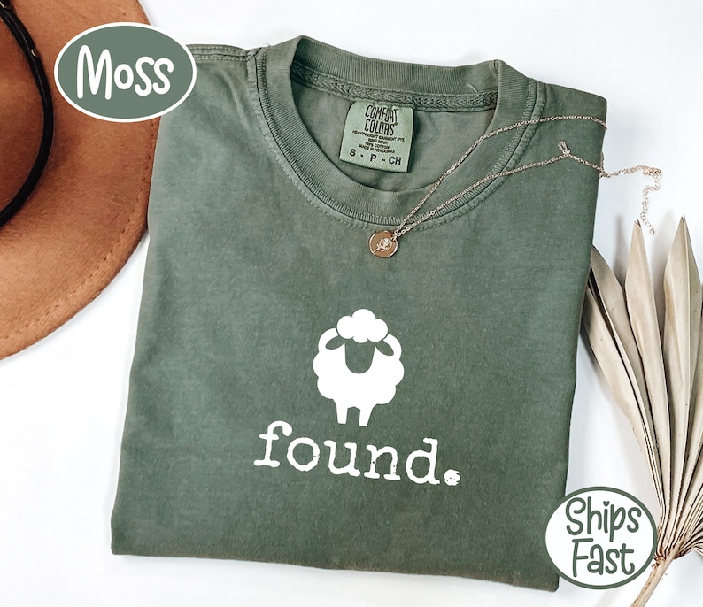 May include: A moss green t-shirt with a white sheep graphic and the word "found" in white. The shirt has a crew neck and short sleeves. A gold necklace with a small pendant is draped on the shirt.
