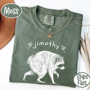May include: A moss green short-sleeve t-shirt featuring a white line-art illustration of a raccoon. The text jimothy is printed in white above the raccoon, flanked by two small heart outlines. The shirt has a Comfort Colors brand neck label.
