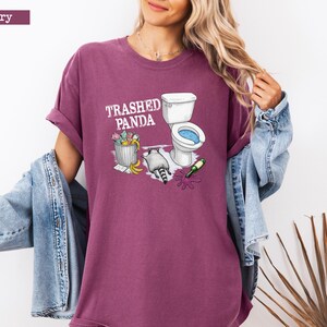 K&ouml;nnte beinhalten: Beerenfarbenes T-Shirt mit dem Text "Trashed Panda" und einer Grafik eines Waschb&auml;ren, einer Toilette und eines M&uuml;lleimers. Der Waschb&auml;r befindet sich neben einer Toilette mit blauem Innenraum. Eine gr&uuml;ne Flasche liegt auf dem Boden.