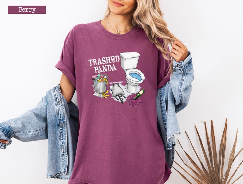 K&ouml;nnte beinhalten: Beerenfarbenes T-Shirt mit dem Text "Trashed Panda" und einer Grafik eines Waschb&auml;ren, einer Toilette und eines M&uuml;lleimers. Der Waschb&auml;r befindet sich neben einer Toilette mit blauem Innenraum. Eine gr&uuml;ne Flasche liegt auf dem Boden.
