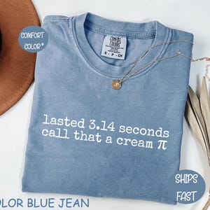 Peut inclure: Un t-shirt à manches courtes de couleur bleu jean avec une encolure ronde. Le t-shirt présente un texte blanc imprimé sur le devant qui indique, lasted 3.14 seconds call that a cream pi. Un petit collier doré avec un pendentif repose dessus.