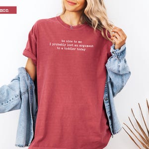 Może przedstawiać: Karmelowa koszulka z białym napisem: "be nice to me, I probably lost an argument to a toddler today". Do tego jasnoniebieska kurtka dżinsowa.