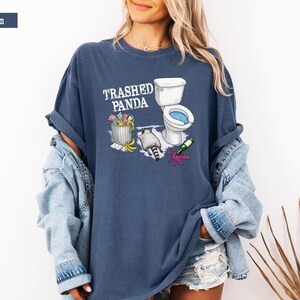 K&ouml;nnte beinhalten: Denimblaues T-Shirt mit dem Text "Trashed Panda" und einer Cartoon-Illustration eines Waschb&auml;ren, einer Toilette und eines M&uuml;lleimers. Der Waschb&auml;r liegt neben einer Toilette und einer versch&uuml;tteten Weinflasche.