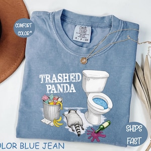 May include: Blue jean coloured t-shirt with the text "Trashed Panda" and a graphic of a raccoon, toilet, bin, and spilled wine. The shirt has a crew neck and short sleeves. A gold necklace is draped on the shirt.