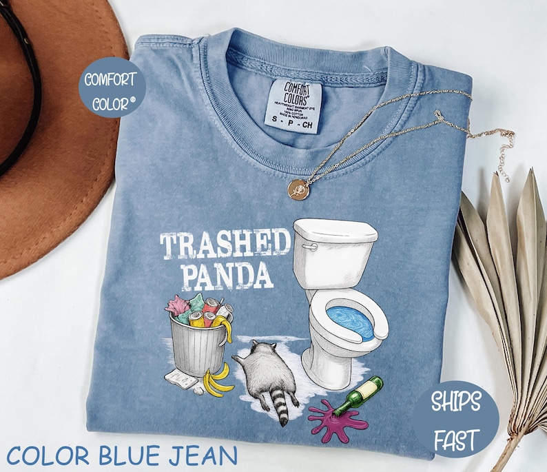 K&ouml;nnte beinhalten: Blaues Jeans-T-Shirt mit dem Text "Trashed Panda" und einer Grafik eines Waschb&auml;ren, einer Toilette, einer M&uuml;lltonne und versch&uuml;ttetem Wein. Das Shirt hat einen Rundhalsausschnitt und kurze &Auml;rmel. Eine goldene Halskette ist auf dem Shirt drapiert.
