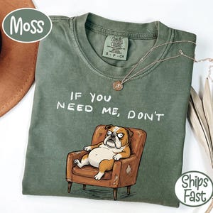 May include: A moss green t-shirt with the text "IF YOU NEED ME, DON'T" and a cartoon bulldog in a brown armchair. The shirt has a crew neck and short sleeves. A gold necklace is draped across the shirt.