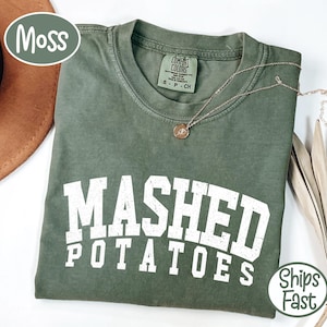 Peut inclure: Un t-shirt vert mousse avec les mots "MASHED POTATOES" en grandes lettres blanches. Un collier en or avec un pendentif repose sur le t-shirt. Le t-shirt est plié et porte une étiquette "Comfort Colors".
