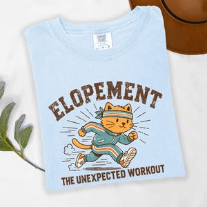 Puede incluir: Camiseta azul claro con las palabras "ELOPEMENT" y "THE UNEXPECTED WORKOUT" en texto marrón. La camiseta presenta un gato de dibujos animados con un chándal y una diadema azules, corriendo con expresión decidida. Es una camiseta de la marca Comfort Colors.