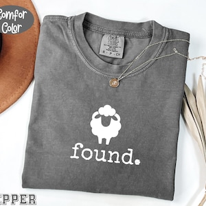 May include: A gray Comfort Colors t-shirt with a white sheep graphic and the word "found." printed below. A silver necklace with a small pendant rests on the shirt. The shirt is folded and displayed with a brown hat and dried plant.