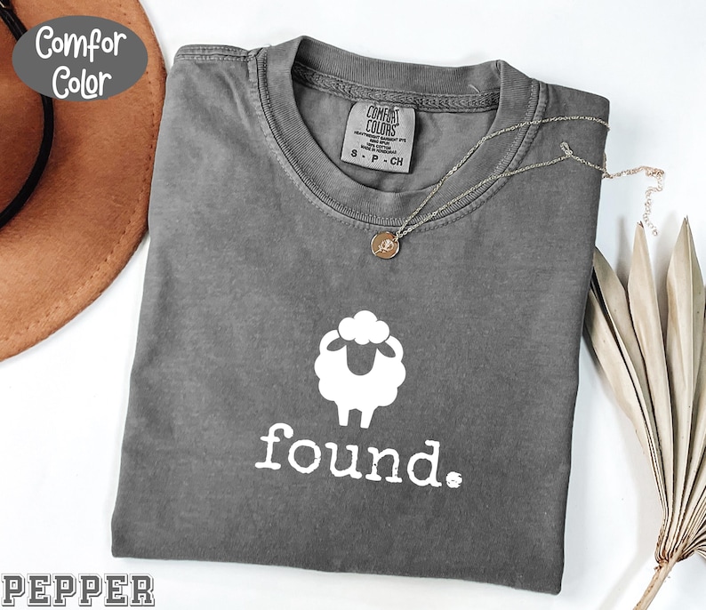 May include: A gray Comfort Colors t-shirt with a white sheep graphic and the word "found." printed below. A silver necklace with a small pendant rests on the shirt. The shirt is folded and displayed with a brown hat and dried plant.