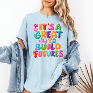 Pode incluir: Camiseta azul clara com a frase "IT'S A GREAT day TO BUILD FUTURES" em letras coloridas. O design inclui flores e uma paleta de cores vibrantes. A camiseta &eacute; feita de um tecido macio e confort&aacute;vel.