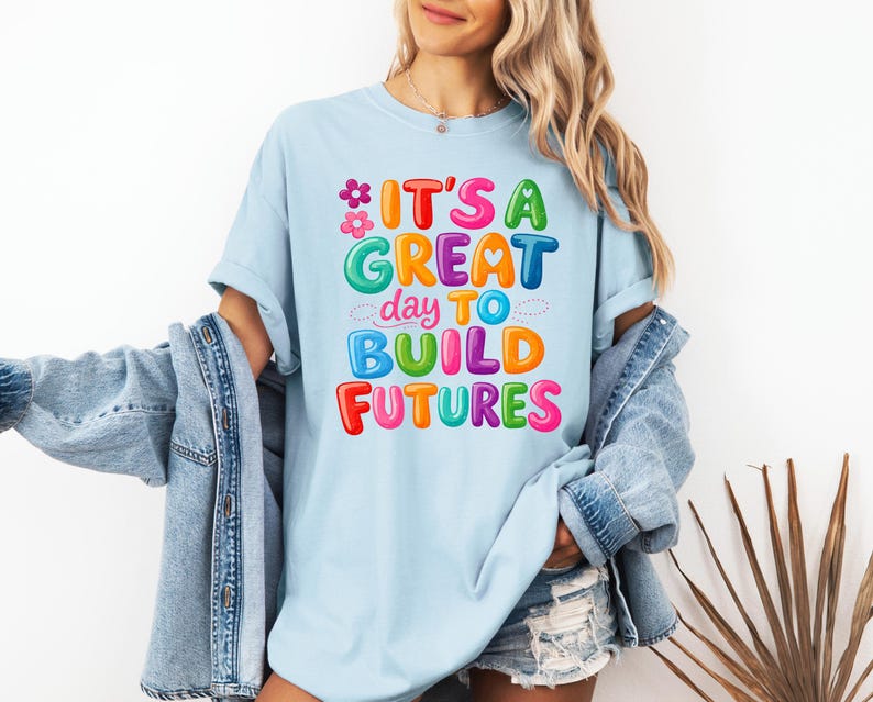 Pode incluir: Camiseta azul clara com a frase "IT'S A GREAT day TO BUILD FUTURES" em letras coloridas. O design inclui flores e uma paleta de cores vibrantes. A camiseta &eacute; feita de um tecido macio e confort&aacute;vel.