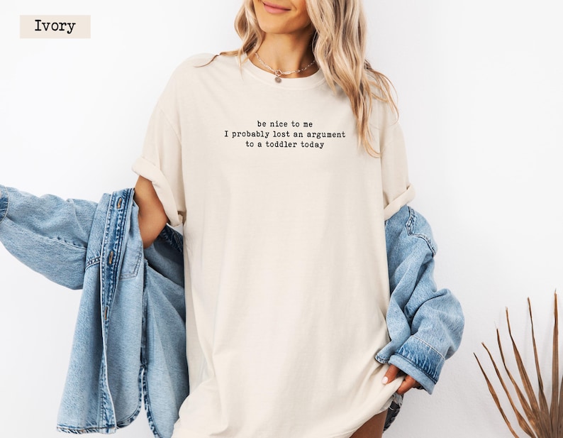 Może przedstawiać: Kremowa koszulka z czarnym napisem "be nice to me I probably lost an argument to a toddler today". Widoczna jest r&oacute;wnież jasnoniebieska kurtka dżinsowa.