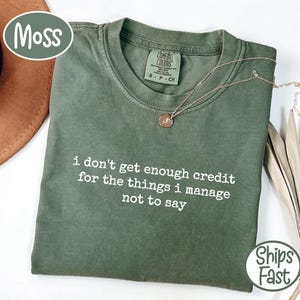 Peut inclure: Un t-shirt vert mousse avec le texte "i don't get enough credit for the things i manage not to say" en blanc. Un collier en or avec un pendentif repose sur le t-shirt. Le t-shirt est plié.
