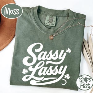 May include: A moss green t-shirt with the words "Sassy Lassy" in white script, accented with shamrocks. A gold necklace with a pendant rests on the shirt. The shirt is folded, and a brown hat is visible in the background.