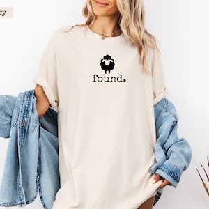 May include: Ivory-colored t-shirt with a black sheep graphic above the word "found." The shirt has a relaxed fit and short sleeves. A light blue denim jacket is draped over the arm.