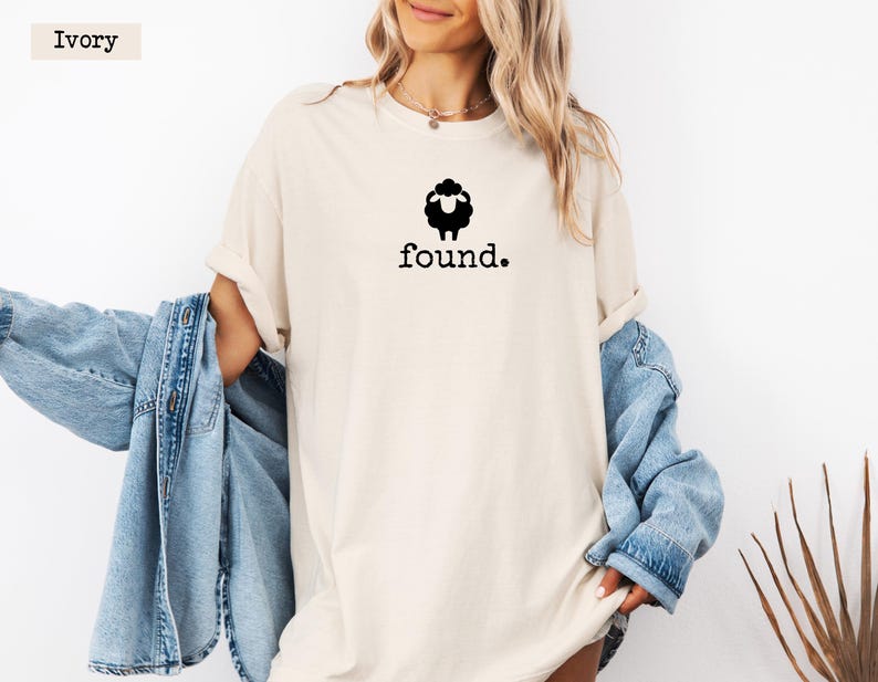May include: Ivory-colored t-shirt with a black sheep graphic above the word "found." The shirt has a relaxed fit and short sleeves. A light blue denim jacket is draped over the arm.