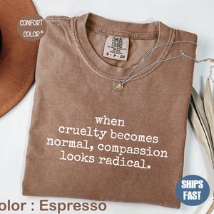 Peut inclure: T-shirt Comfort Colors couleur Espresso avec la citation blanche "when cruelty becomes normal, compassion looks radical". Un collier argenté avec un petit pendentif est sur le t-shirt. Taille S-P-CH.