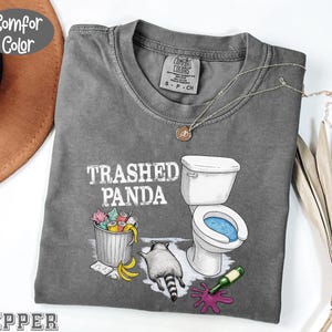 K&ouml;nnte beinhalten: Ein graues Comfort Colors T-Shirt mit dem Text "Trashed Panda" und einer Grafik eines Waschb&auml;ren, einer Toilette und eines M&uuml;lleimers. Das Shirt hat einen Rundhalsausschnitt und kurze &Auml;rmel. Eine silberne Halskette mit Anh&auml;nger befindet sich auf dem Shirt.