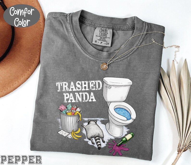 K&ouml;nnte beinhalten: Ein graues Comfort Colors T-Shirt mit dem Text "Trashed Panda" und einer Grafik eines Waschb&auml;ren, einer Toilette und eines M&uuml;lleimers. Das Shirt hat einen Rundhalsausschnitt und kurze &Auml;rmel. Eine silberne Halskette mit Anh&auml;nger befindet sich auf dem Shirt.