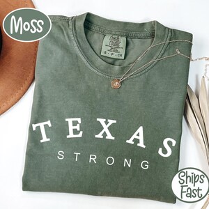 Peut inclure: Un t-shirt vert mousse avec les mots "TEXAS STRONG" en lettres blanches. Un collier en or avec un pendentif repose sur le t-shirt. Le t-shirt est plié et présenté avec un chapeau marron et une plante séchée.