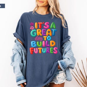 Pode incluir: Camiseta azul denim com a frase "IT'S A GREAT day TO BUILD FUTURES" em letras coloridas. A camiseta &eacute; usada com uma jaqueta jeans e shorts. O estilo geral &eacute; casual e otimista.