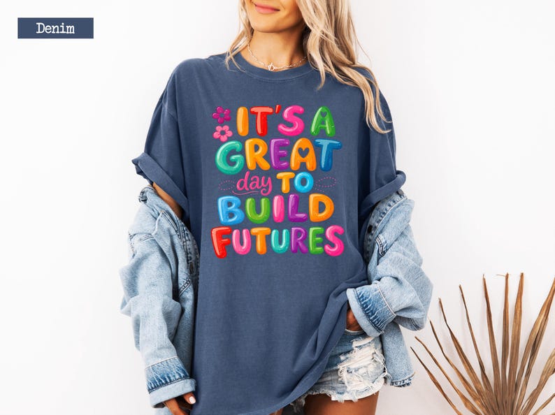 Pode incluir: Camiseta azul denim com a frase "IT'S A GREAT day TO BUILD FUTURES" em letras coloridas. A camiseta &eacute; usada com uma jaqueta jeans e shorts. O estilo geral &eacute; casual e otimista.