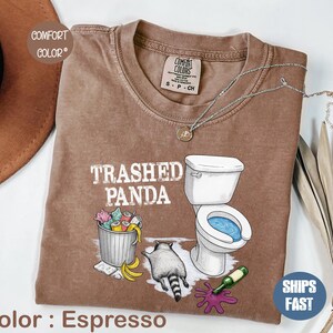 K&ouml;nnte beinhalten: Espressofarbenes T-Shirt mit dem Text "Trashed Panda" und einer Grafik eines Waschb&auml;ren, einer Toilette und einer M&uuml;lltonne. Das Shirt hat einen Rundhalsausschnitt und kurze &Auml;rmel. Eine silberne Halskette ist &uuml;ber dem Shirt drapiert.