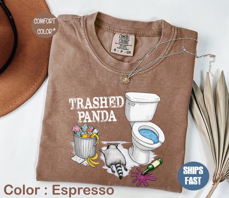 K&ouml;nnte beinhalten: Espressofarbenes T-Shirt mit dem Text "Trashed Panda" und einer Grafik eines Waschb&auml;ren, einer Toilette und einer M&uuml;lltonne. Das Shirt hat einen Rundhalsausschnitt und kurze &Auml;rmel. Eine silberne Halskette ist &uuml;ber dem Shirt drapiert.