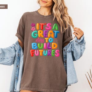 Pode incluir: Camiseta cor de caf&eacute; com a frase "IT'S A GREAT DAY TO BUILD FUTURES" em letras coloridas de bolhas. A camiseta tem mangas enroladas e &eacute; usada com uma jaqueta jeans azul.